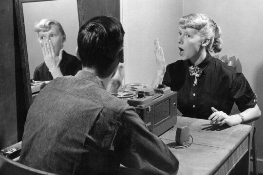 Person holding hand in front of face with an open-mouthed expression looking at another person sitting across table.