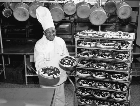 Chef Carson Gulley in kitchen holding two fudge-bottom pies standing beside bakery rack lined with more pies.