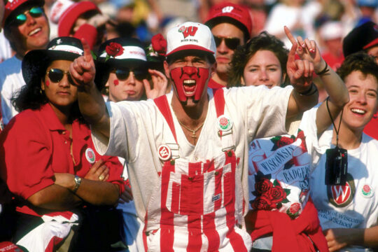 Spirited Badger fan in crowd at football game wearing red and white striped coveralls with painted face boasting we're number one.